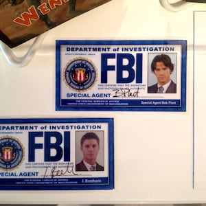 May include: Two blue and white FBI identification cards with the text "Department of Investigation" and "FBI" in bold, white letters. The cards feature a seal with a shield and the text "Special Agent" and the signature of the agent. The cards are on a white surface with text about the show Supernatural.