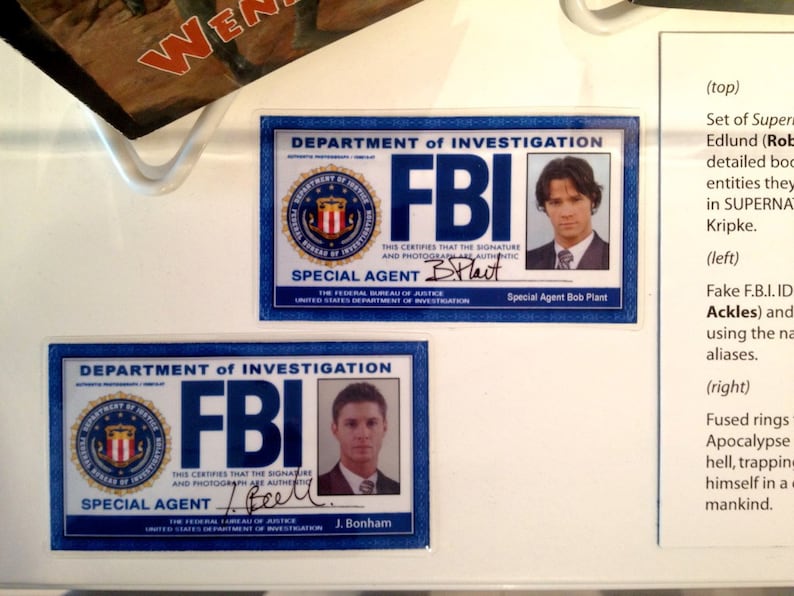 May include: Two blue and white FBI identification cards with the text "Department of Investigation" and "FBI" in bold, white letters. The cards feature a seal with a shield and the text "Special Agent" and the signature of the agent. The cards are on a white surface with text about the show Supernatural.