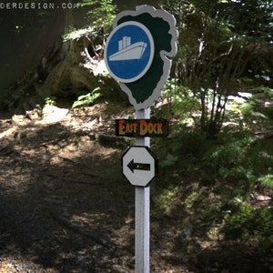 May include: A wooden signpost with three signs. The top sign is blue with a white ship and the text "PAUL ELDER DESIGN.COM". The middle sign is black with white text that says "EAST DOCK". The bottom sign is white with a black arrow pointing to the left.