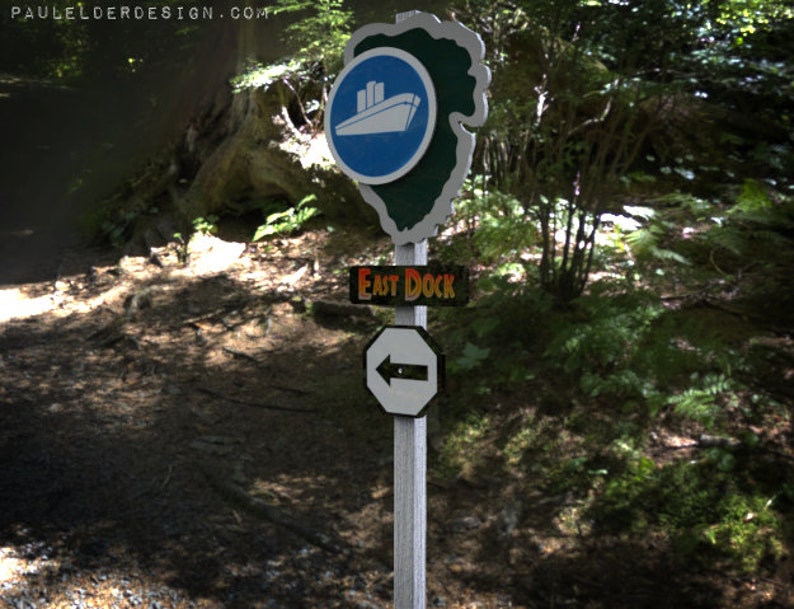 May include: A wooden signpost with three signs. The top sign is blue with a white ship and the text "PAUL ELDER DESIGN.COM". The middle sign is black with white text that says "EAST DOCK". The bottom sign is white with a black arrow pointing to the left.