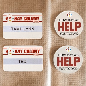 Può includere: Due cartellini nominativi bianchi con il testo "BAY COLONY FOODS" e i nomi "TAMI-LYNN" e "TED" stampati su di essi. Due pulsanti rotondi bianchi con il testo "HOW MAY WE HELP YOU TODAY?" stampato su di essi.