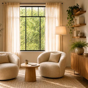 May include: Two cream-colored, rounded, textured fabric armchairs with matching throw pillows are positioned on a woven rug. Between them sits a round wooden side table with a ceramic mug and a book. A floor lamp stands nearby.