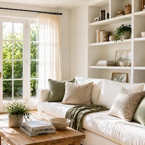 May include: A light and spacious living room featuring a white sofa with green and beige cushions and a green throw. A wooden coffee table displays books and a bowl. A white bookshelf and sheer curtains are also visible, creating a relaxed atmosphere.