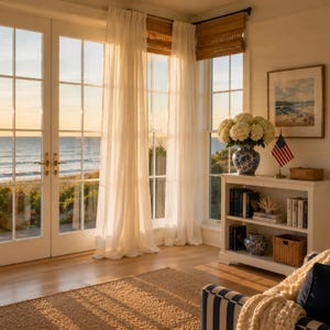 Puede incluir: Una habitación soleada con vistas al océano a través de grandes ventanas. Cortinas blancas transparentes enmarcan las ventanas, y una alfombra tejida se encuentra en el suelo de madera. Una estantería blanca contiene libros y objetos decorativos.