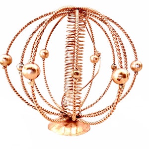Puede incluir: Una escultura de alambre de cobre que representa una esfera con una espiral en el centro y esferas más pequeñas unidas a los anillos exteriores.