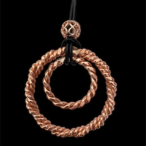 May include: A rose gold necklace with a double circle pendant. The circles are made of twisted wire and are connected to a black leather cord.