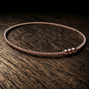 May include: A rose gold bangle bracelet with a twisted rope design. The bracelet features three small, round beads and rests on a wooden surface. The jewelry is a simple, elegant accessory.