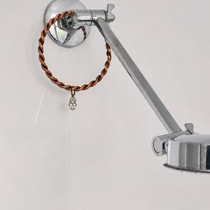 May include: A chrome-plated metal towel ring with a twisted copper-coloured circular bar. A small silver-toned Hamsa hand charm hangs from the bottom of the twisted ring. The towel ring is mounted on a circular chrome base attached to a wall.
