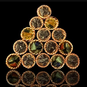 Op de afbeelding: Een driehoekige opstelling van 15 kleine, ronde, koperkleurige containers met gouden versieringen. Elke container is gevuld met een verscheidenheid aan kleurrijke stenen en kristallen.