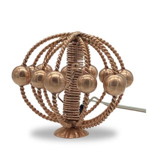 Puede incluir: Objeto decorativo de color cobre con diseño esférico. Presenta una estructura central en espiral, rodeada de anillos de alambre trenzado y pequeñas esferas. La base es un pequeño pedestal redondo.