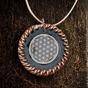 May include: A circular pendant with a black central design, displaying a white geometric pattern, framed by a copper-toned, rope-style edge. It is suspended from a white cord, set against a dark, textured backdrop.