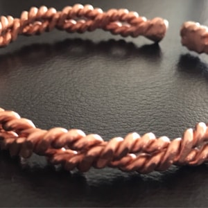 Op de afbeelding: Een koperkleurige, open manchetarmband met een gedraaid touwontwerp. De armband is gemaakt van in elkaar verweven strengen, waardoor een textuur ontstaat. De sieraden worden gepresenteerd op een donkere ondergrond.