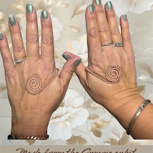 Puede incluir: Dos manos que muestran espirales de mano tensoras de cobre. Las manos tienen esmalte de uñas verde brillante, anillos y pulseras. El texto en la parte inferior dice "Made from the Gympie cubit Tensor hand Spirals."