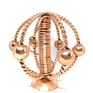 Pode incluir: Uma escultura de metal de cor cobre com um design em espiral e cinco bolas redondas. A escultura está sobre uma base e tem um design de fio torcido.