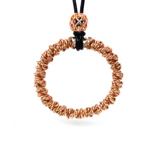 Puede incluir: Un collar dorado con un colgante circular hecho de alambre retorcido. El colgante está unido a un cordón de cuero negro con una cuenta decorativa en la parte superior.