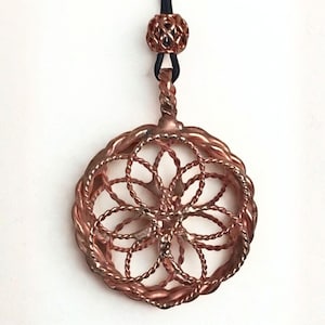Könnte beinhalten: Kupferfarbener Anhänger mit kreisförmigem, durchbrochenem Design. Der Anhänger hat ein zentrales Blumenmuster, das von einem geflochtenen Rand umgeben ist. Er hängt an einer schwarzen Kordel und hat eine dekorative Perle oben. Wahrscheinlich ein Schmuckstück.
