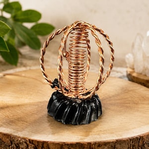 Puede incluir: Un objeto decorativo que presenta una bobina central de alambre de cobre rodeada por dos anillos entrelazados de alambre de cobre trenzado. La estructura está montada sobre una base negra, circular y estriada. El cobre tiene un acabado metálico.