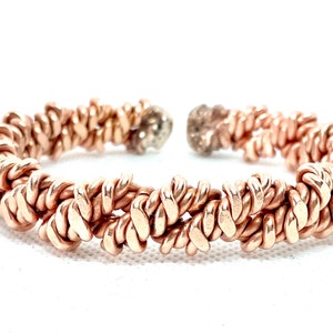 May include: A rose gold braided cuff bracelet. The bracelet is made of a series of interwoven metal loops.