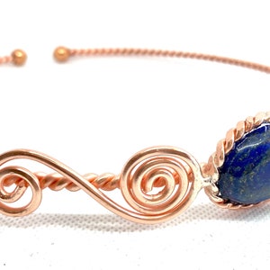 May include: A rose gold wire bracelet with a blue lapis lazuli stone. The bracelet has a twisted wire design and two spiral accents.