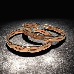 May include: Three gold-coloured, open-ended rings with a braided design. The rings are displayed on a dark, textured surface, with the light creating shadows that highlight the texture and shape of the jewellery.