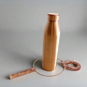 May include: A copper water bottle with a cleaning brush. The bottle is tall and slender with a rounded bottom and a smooth, shiny surface. The cleaning brush is made of copper wire and has a long, thin handle.