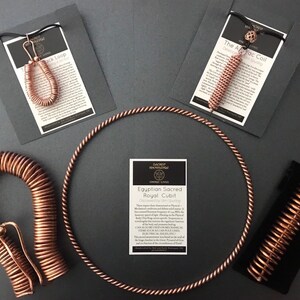 May include: A collection of copper wire sculptures, including a circle, a coil, a loop, and a curved shape. The sculptures are displayed on a grey surface with cards that describe them. The cards include the text "The Aztec Coil", "The Lock Loop", and "Egyptian Sacred Royal Cubit".