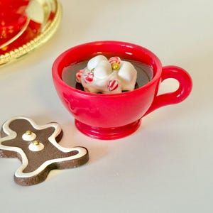 May include: A festive scene featuring a red mug filled with a dark liquid and topped with marshmallows and candy canes. A gingerbread man cookie and a gold-rimmed plate are also present, creating a holiday theme.