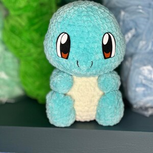Pu&ograve; includere: Un giocattolo di peluche blu e bianco all'uncinetto che ricorda una tartaruga dei cartoni animati con occhi grandi. Il giocattolo &egrave; seduto su uno scaffale blu scuro.