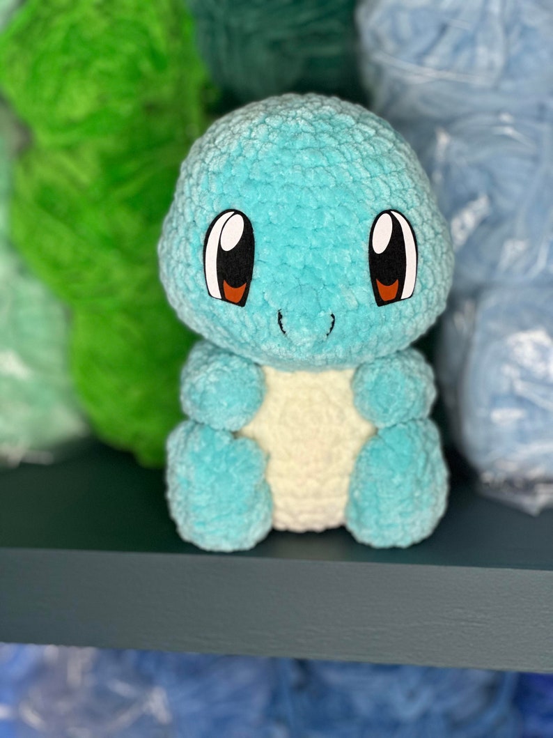 Pu&ograve; includere: Un giocattolo di peluche blu e bianco all'uncinetto che ricorda una tartaruga dei cartoni animati con occhi grandi. Il giocattolo &egrave; seduto su uno scaffale blu scuro.