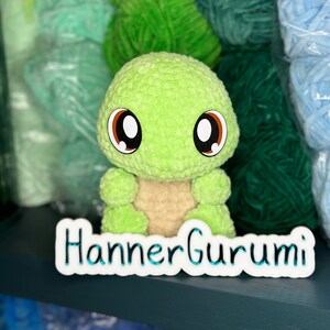 Pu&ograve; includere: Un giocattolo amigurumi a forma di tartaruga verde all'uncinetto con grandi occhi marroni. Il giocattolo &egrave; seduto su uno scaffale con gomitoli di lana in varie tonalit&agrave; di verde e blu. Lo scaffale &egrave; etichettato "Hanner Gurumi".