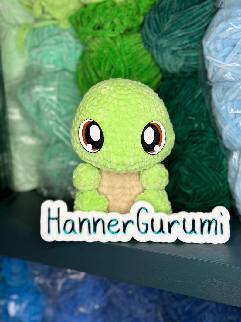 Pu&ograve; includere: Un giocattolo amigurumi a forma di tartaruga verde all'uncinetto con grandi occhi marroni. Il giocattolo &egrave; seduto su uno scaffale con gomitoli di lana in varie tonalit&agrave; di verde e blu. Lo scaffale &egrave; etichettato "Hanner Gurumi".