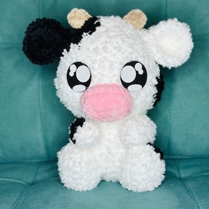 May include: A white and black crocheted plush cow toy with large black eyes and a pink nose. The cow is sitting on a teal green surface.
