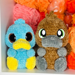 Può includere: Due animali di peluche all'uncinetto, un ornitorinco blu e un ornitorinco marrone, con grandi occhi rotondi e sporgenti. Gli ornitorinchi sono seduti su una superficie con filato sullo sfondo.
