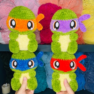 Pu&ograve; includere: Quattro peluche di Tartarughe Ninja Adolescenti all'uncinetto. Ogni tartaruga &egrave; verde con una maschera di colore diverso: arancione, viola, blu e rosso. Le tartarughe stanno tenendo le mani davanti a loro.