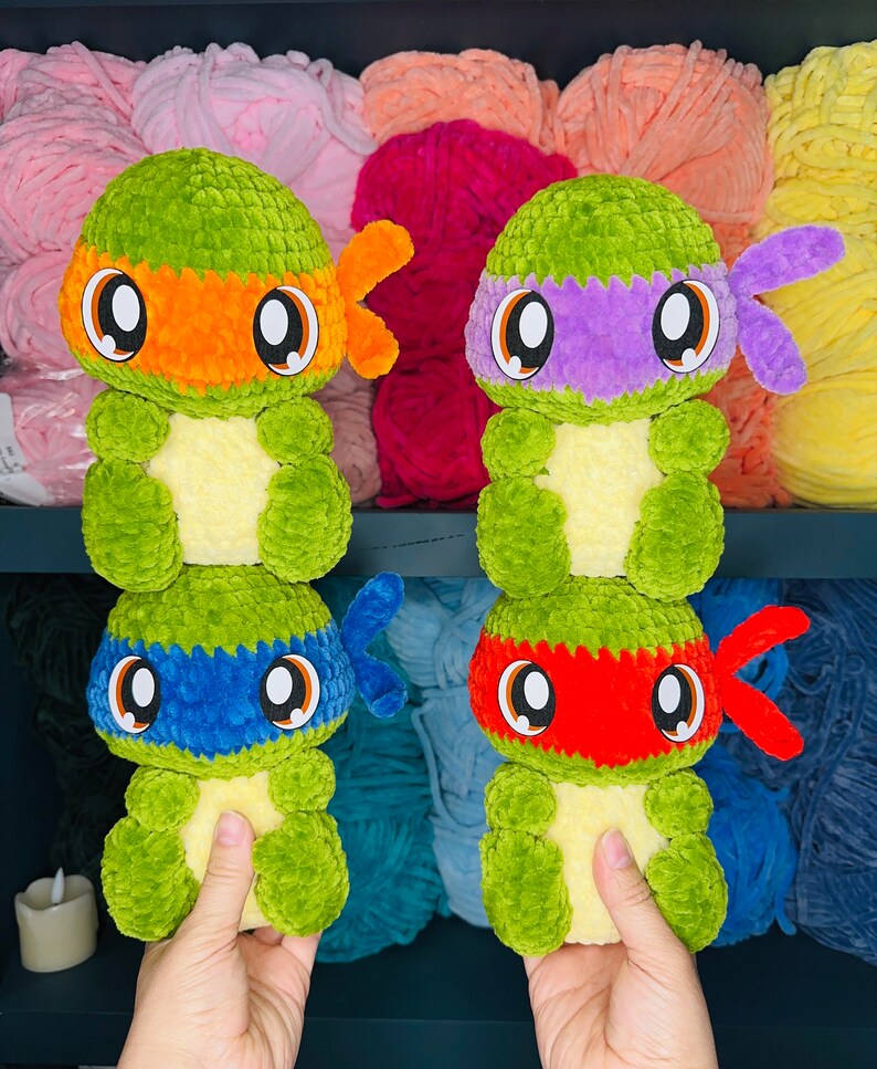 Pu&ograve; includere: Quattro peluche di Tartarughe Ninja Adolescenti all'uncinetto. Ogni tartaruga &egrave; verde con una maschera di colore diverso: arancione, viola, blu e rosso. Le tartarughe stanno tenendo le mani davanti a loro.