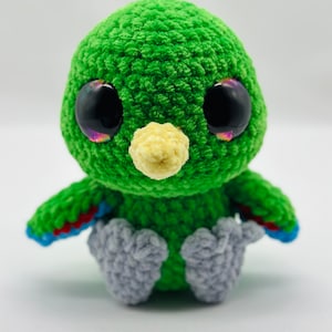 May include: A green crocheted bird with large black eyes and a yellow beak. The bird is sitting on a white surface.