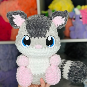 Puede incluir: Un juguete de peluche tejido a crochet en forma de animal gris y blanco con orejas y patas rosas. El juguete tiene grandes ojos azules y una nariz rosa.