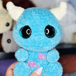 May include: A blue crocheted plush toy with white horns and large black eyes. The toy has a pink and white pattern on its body.