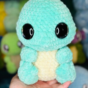 Pu&ograve; includere: Un giocattolo di peluche all'uncinetto a forma di piccola creatura verde con grandi occhi neri. Il giocattolo ha una pancia giallo chiaro ed &egrave; seduto nella mano di una persona.