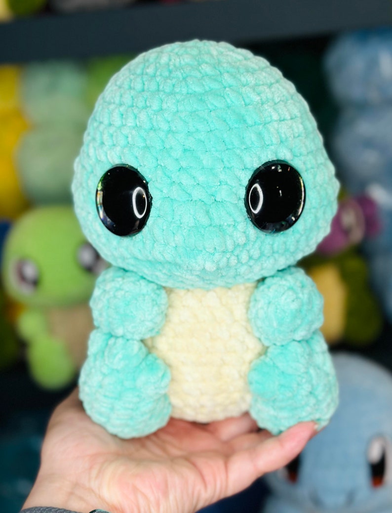Pu&ograve; includere: Un giocattolo di peluche all'uncinetto a forma di piccola creatura verde con grandi occhi neri. Il giocattolo ha una pancia giallo chiaro ed &egrave; seduto nella mano di una persona.
