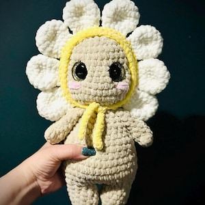 May include: A handmade crochet doll in the shape of a sunflower. The doll has a beige body, large black eyes, and a yellow hood. The sunflower petals are white. The doll is soft and plush.