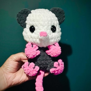 May include: A handmade crochet plush toy of an opossum featuring a white head, dark grey ears, and a dark grey body. The creature has large black plastic eyes, a small pink nose, and bright pink hands, feet, and a long, curled tail made of yarn.