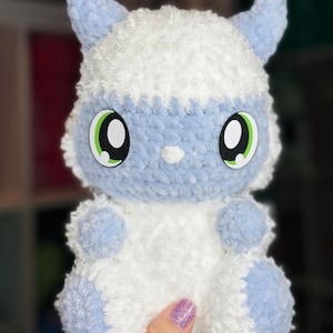 May include: A handmade plush toy in the shape of a cartoon creature. The toy is primarily white with light blue accents on the face, ears, and paws. It has large, cartoonish eyes with black pupils and green highlights. The plush is held in a hand.