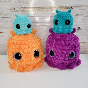 Peut inclure: Deux jouets en peluche crochetés avec des yeux globuleux. Le plus grand jouet est violet et le plus petit est bleu sarcelle. Le plus petit jouet est assis sur un plus grand jouet en peluche crochetée orange avec des yeux globuleux.