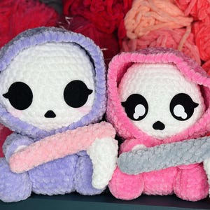 Könnte beinhalten: Zwei gehäkelte Plüschtiere in Form von Skeletten, die Kapuzenoutfits tragen. Das Skelett links trägt ein lilafarbenes Kapuzenoutfit und hält eine rosa gehäkelte Sense. Das Skelett rechts trägt ein rosafarbenes Kapuzenoutfit und hält eine graue gehäkelte Sense.