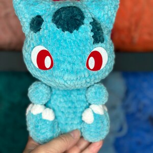 Pu&ograve; includere: Un giocattolo di peluche blu all'uncinetto che ricorda un personaggio di Pokemon con occhi rossi e piedi bianchi.