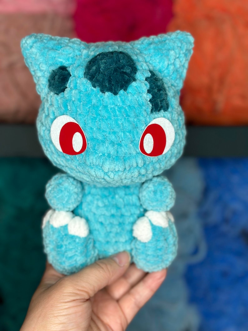 Pu&ograve; includere: Un giocattolo di peluche blu all'uncinetto che ricorda un personaggio di Pokemon con occhi rossi e piedi bianchi.