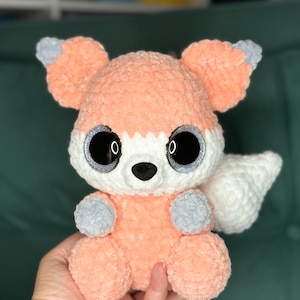 May include: A crocheted fox plush toy with a peach-coloured body, white tail, and grey accents. The toy has large black eyes with glitter accents.