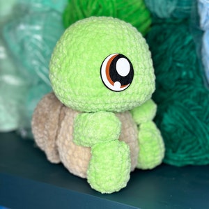 Pu&ograve; includere: Un giocattolo di peluche di tartaruga verde all'uncinetto con occhi marroni e bianchi. Il giocattolo &egrave; seduto su uno scaffale blu scuro.