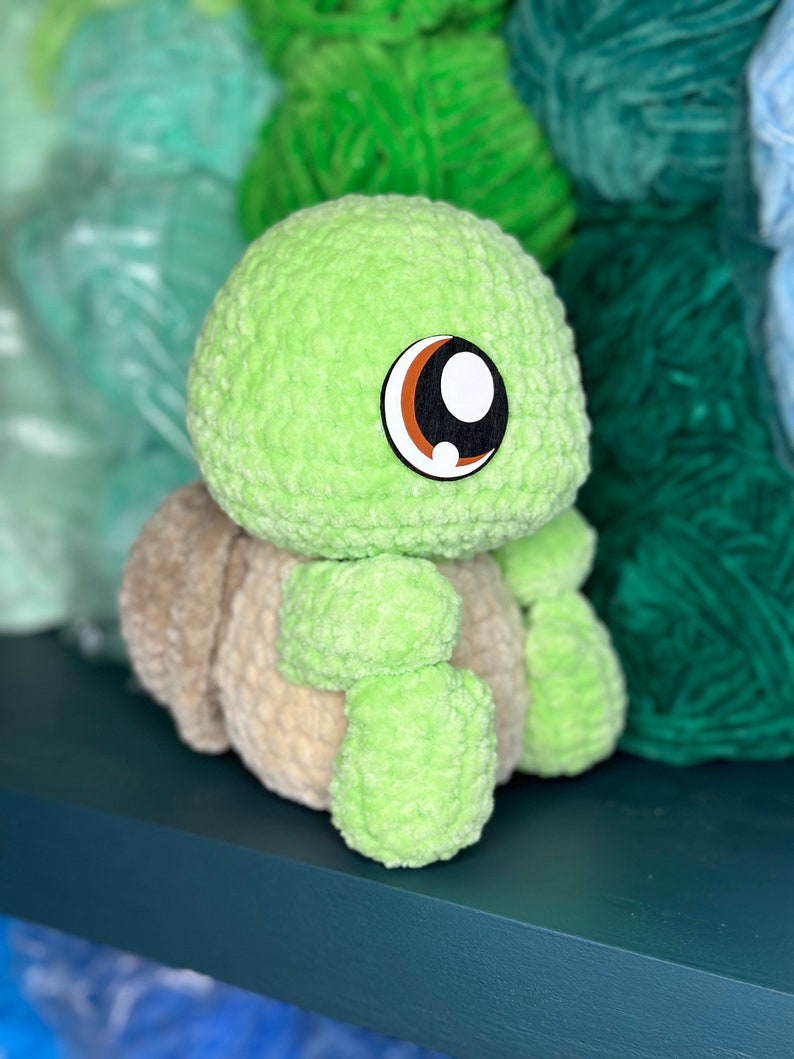 Pu&ograve; includere: Un giocattolo di peluche di tartaruga verde all'uncinetto con occhi marroni e bianchi. Il giocattolo &egrave; seduto su uno scaffale blu scuro.